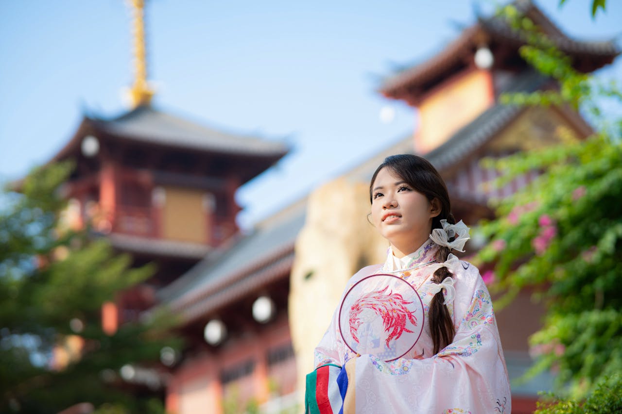 A young woman in traditional Asian attire poses by a colorful pagoda in a sunny outdoor setting.