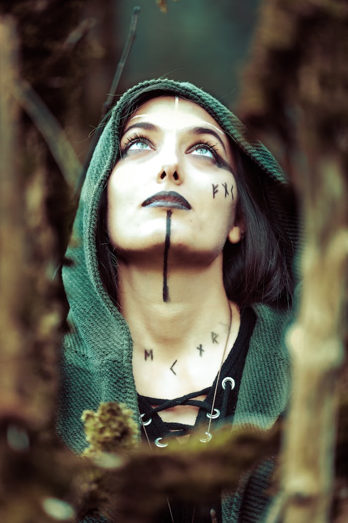 A woman in a green hood with rune symbols looks upwards amidst a forest setting.