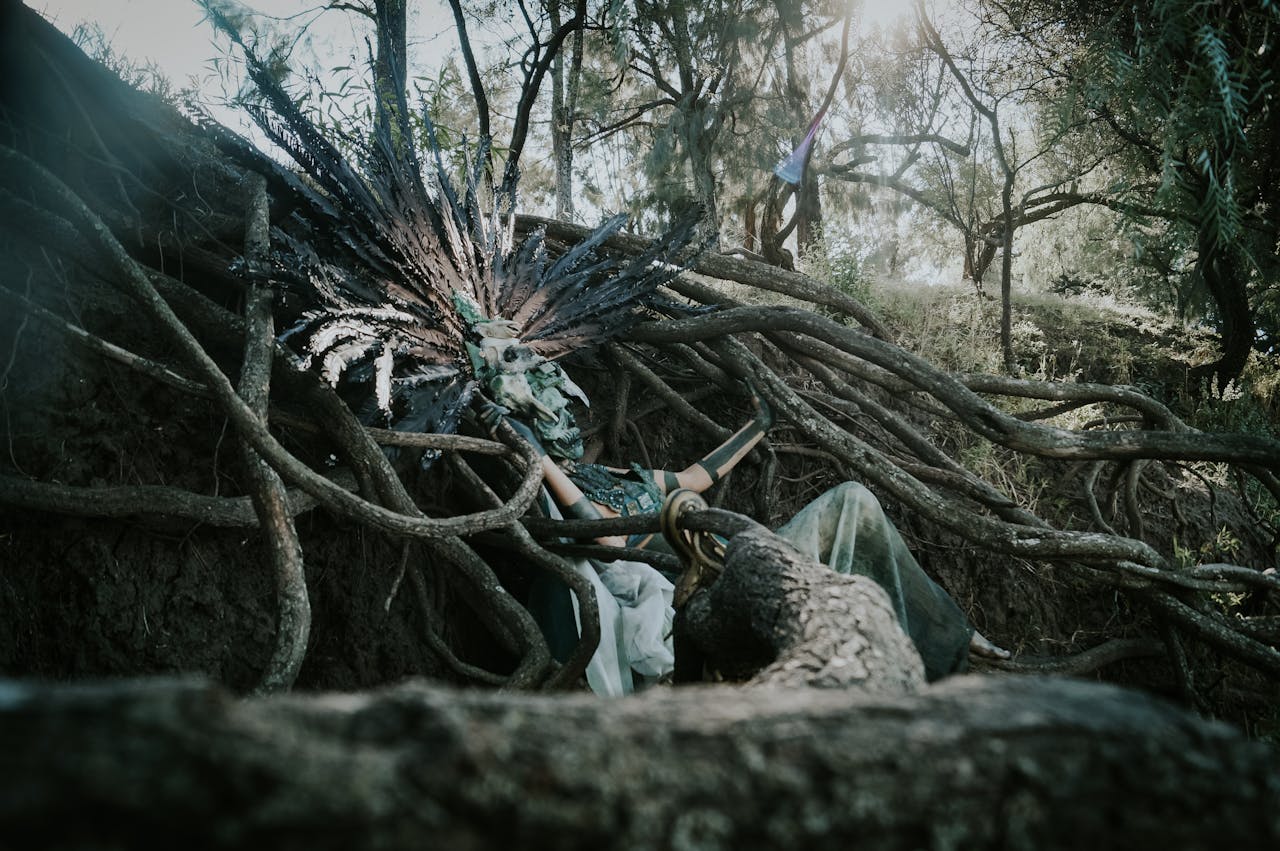 A mysterious figure in elaborate costume rests among twisted tree roots in a forest.